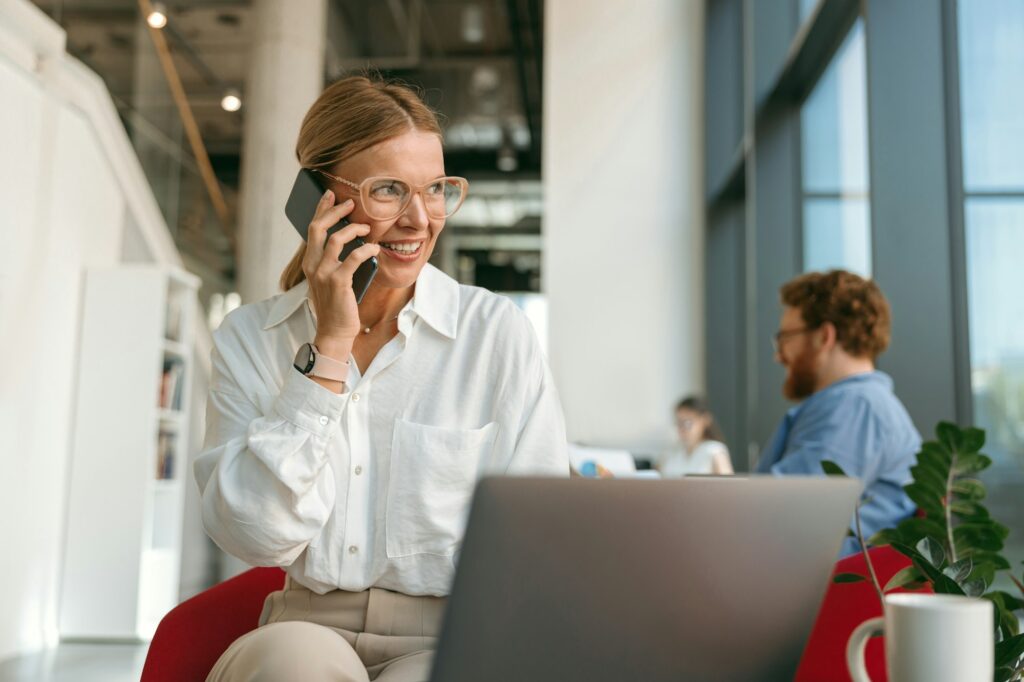 Smiling woman freelancer talking on phone with client while working on laptop in modern coworking