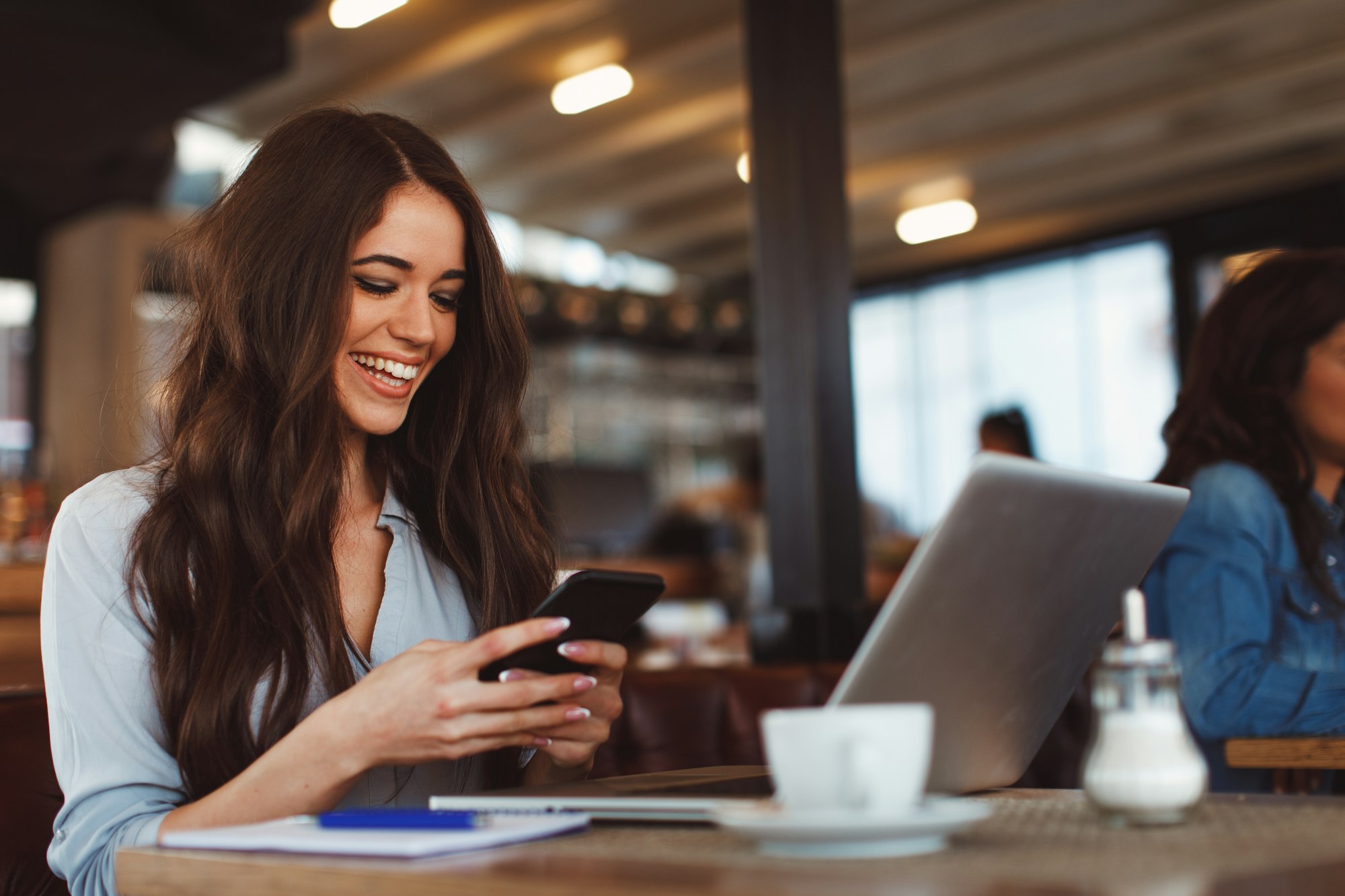 Young woman texting on the phone with a laptop on table in cafe